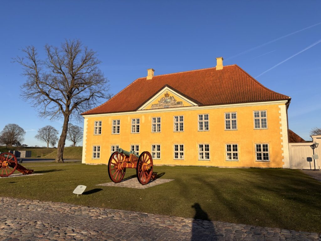Gul bygning med rødt tegltag inde på Kastellet med to gamle kanoner på plænen.